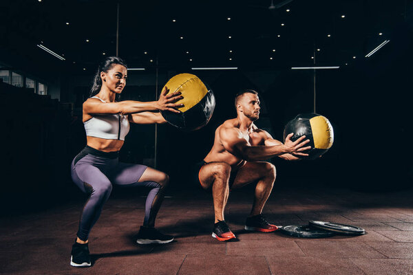 атлетический спортсмен и спортсменка делают приседания с мячами медицины вместе в темном зале
