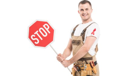 happy adult builder with aids awareness red ribbon on overall holding stop road sign and looking at camera isolated on white clipart