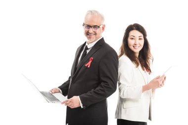 multiethnic happy businesspeople with aids awareness red ribbons working with gadgets while standing back to back and looking at camera isolated on white clipart