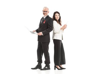 successful adult businesspeople with aids awareness red ribbons working with gadgets while standing back to back and looking at camera isolated on white clipart