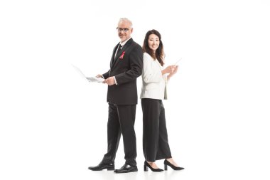happy adult businesspeople with aids awareness red ribbons working with gadgets while standing back to back and looking at camera isolated on white clipart