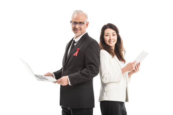 smiling adult businesspeople with aids awareness red ribbons working with gadgets while standing back to back and looking at camera isolated on white