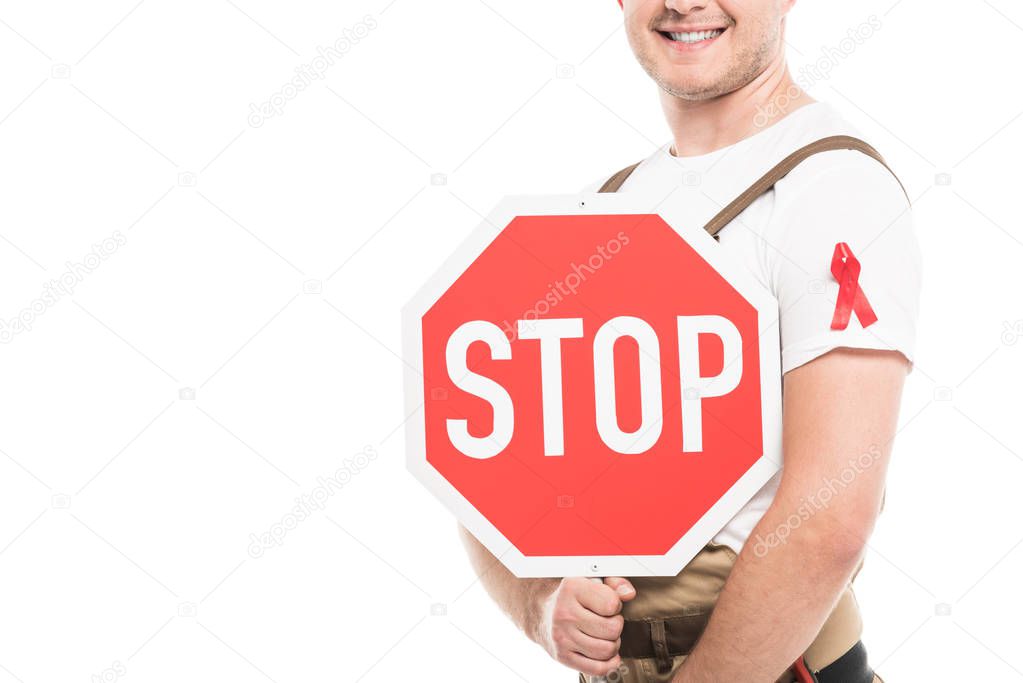 cropped shot of smiling builder with aids awareness red ribbon on overall holding stop road sign isolated on white