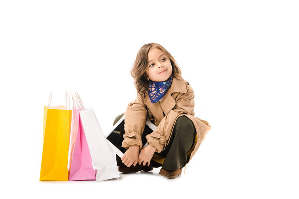 beautiful little child in trench coat sitting on floor with colorful shopping bags isolated on white
