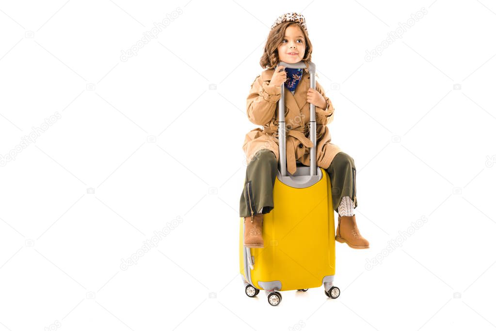 adorable little child in trench coat sitting on yellow suitcase isolated on white