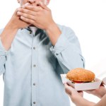 Cropped image of man covering mouth, woman proposing burger isolated on white