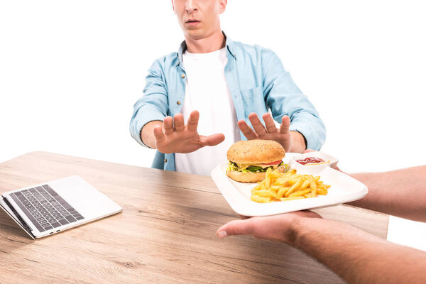 cropped image of man rejecting unhealthy burger and french fries at table isolated on white