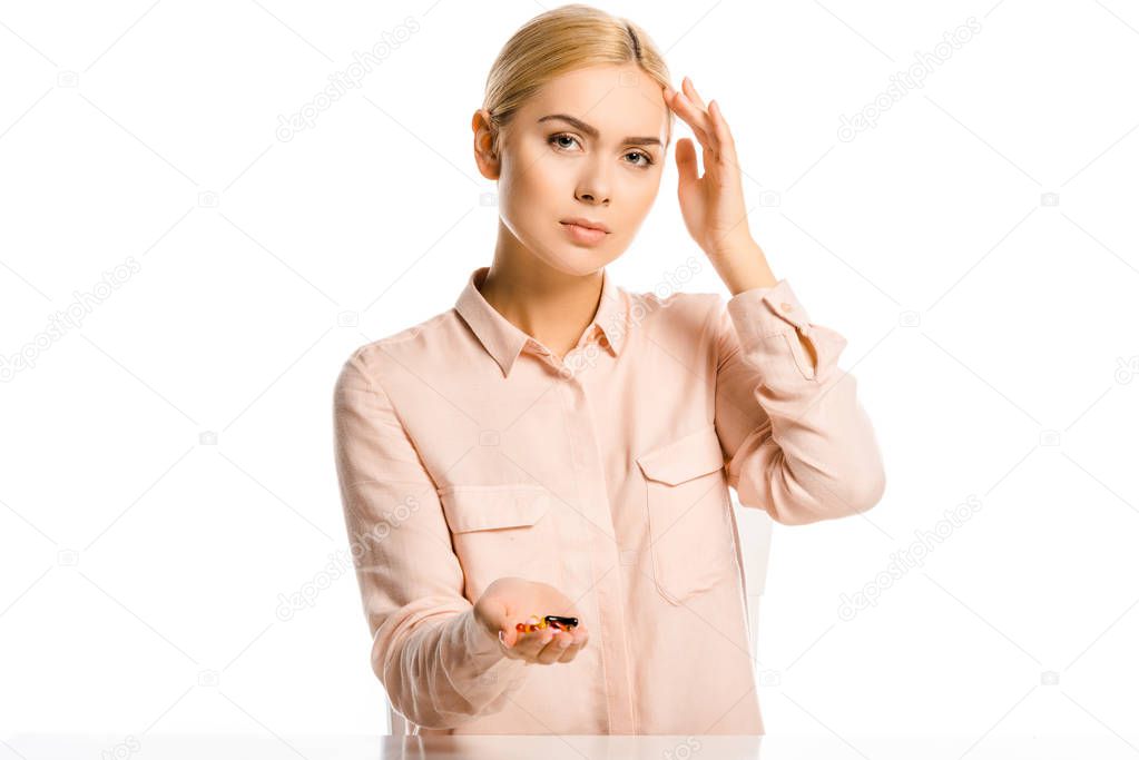 attractive woman holding pills, touching head and looking at camera isolated on white