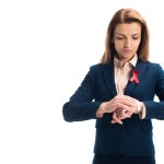 Attractive businesswoman with red ribbon on suit checking time isolated on white, world aids day concept