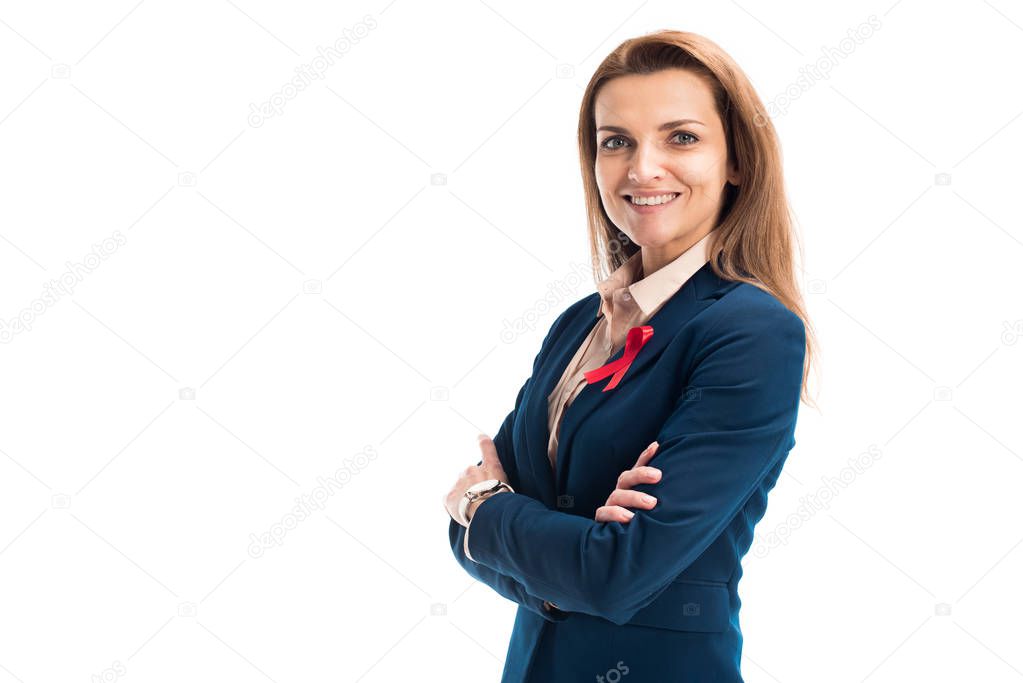 smiling attractive businesswoman with red ribbon on suit standing with crossed arms isolated on white, world aids day concept