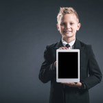 Porträt eines lächelnden Jungen, der wie ein Geschäftsmann gekleidet ist und Tablet mit leerem Bildschirm in der Hand vor grauem Hintergrund zeigt