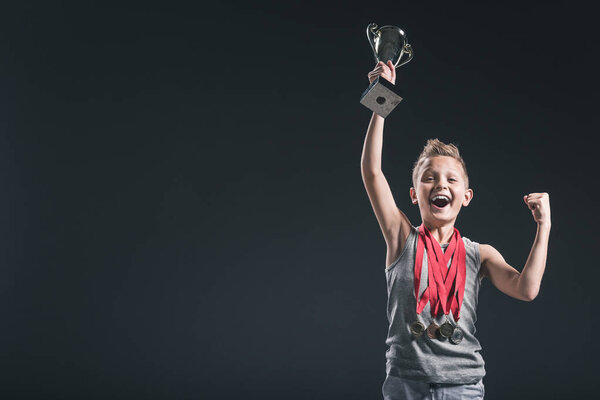 портрет счастливого мальчика в спортивной одежде с медалями и кубками чемпионов жестом на черном фоне
