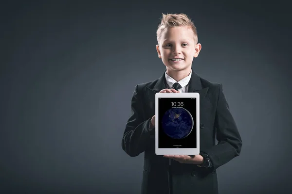灰色の背景に手でタブレットを示す実業家のような服を着て微笑む少年の肖像画 — ストック写真