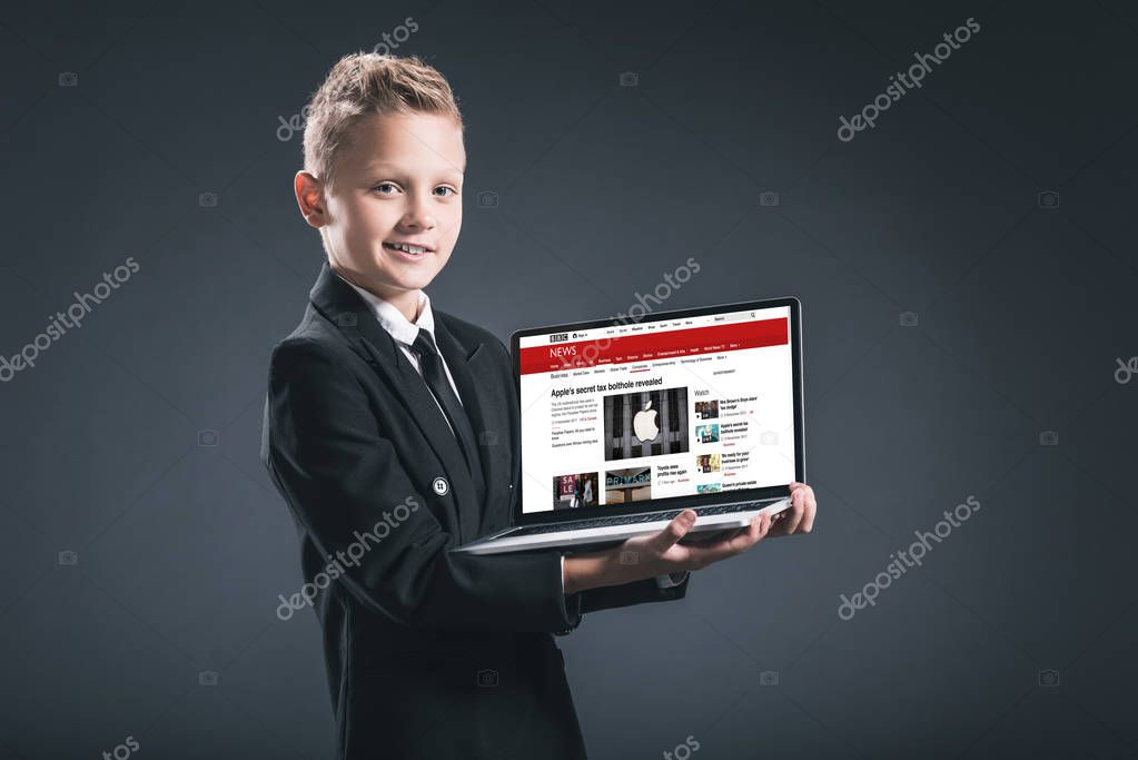 Smiling boy in businessman suit showing laptop with bbc website on grey background
