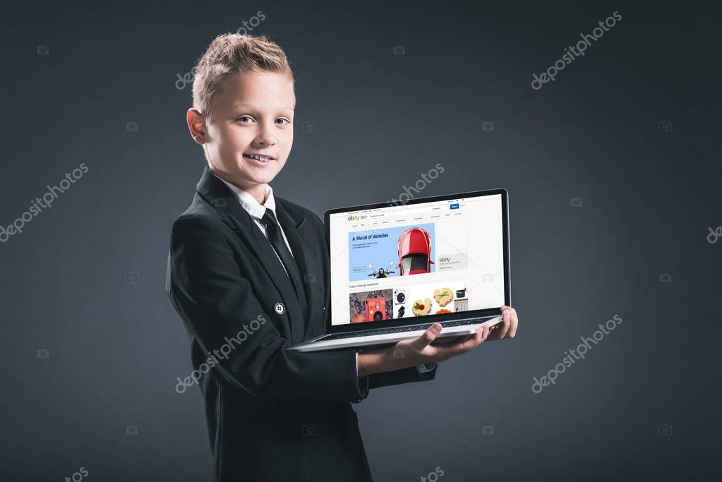 Smiling boy in businessman suit showing laptop with ebay website on grey background