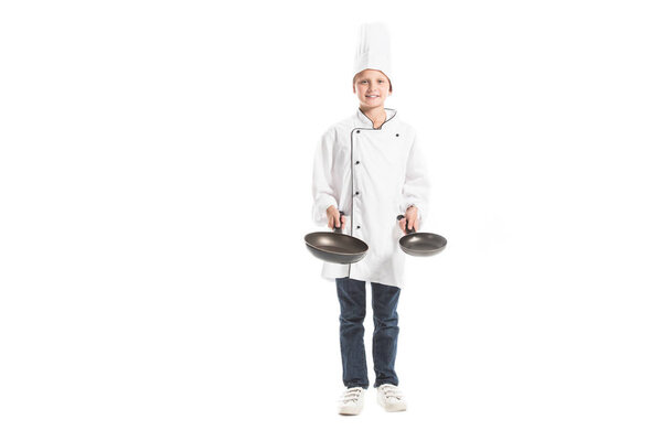smiling boy in white chef uniform and hat holding frying pans and looking at camera isolated on white