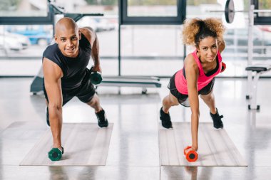 cheerful african american couple of athletes exercising with dumbbells on fitness mats at gym clipart