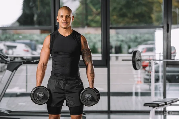 微笑的非洲裔美国运动员看相机和锻炼哑铃在健身房 — 图库照片