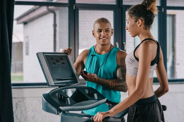 微笑的非洲裔美国男性教练与定时器看女运动员运行在跑步机在健身房 — 图库照片