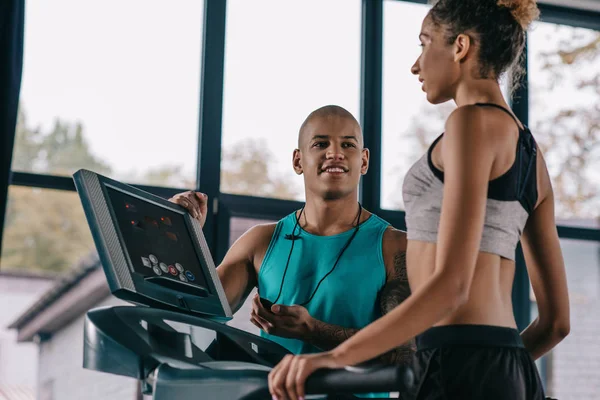 年轻的男性教练与定时器看女运动员运行在跑步机在健身房 — 免费的图库照片