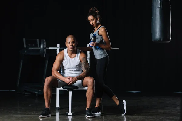美丽的非洲裔美国人女运动员与运动瓶站立附近的男朋友在健身房 — 图库照片