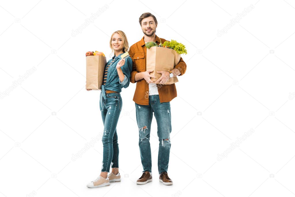 attractive cheerful girl showing credit card while her boyfriend standing near with shopping bags full of products isolated on white