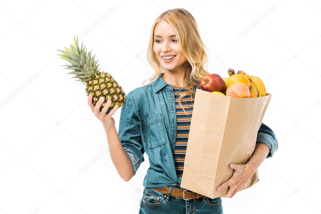 cheerful woman looking at pineapple and holding paper bag with products isolated on white