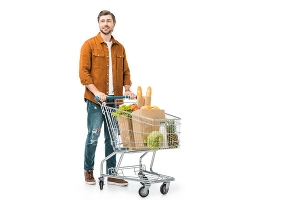 cheerful man carrying shopping trolley with products in paper bags isolated on white