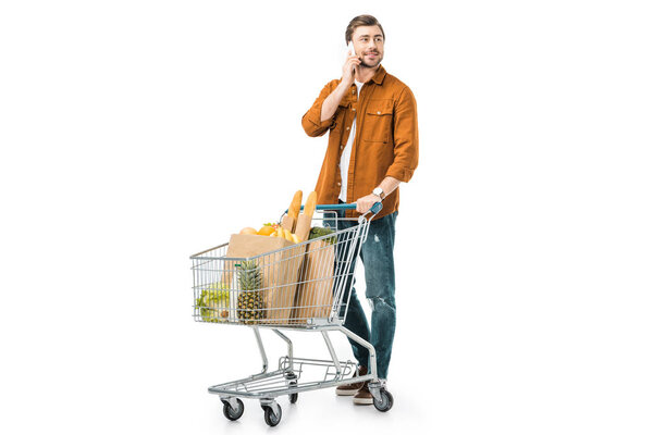 cheerful man carrying shopping trolley with products in paper bags and talking on smartphone isolated on white