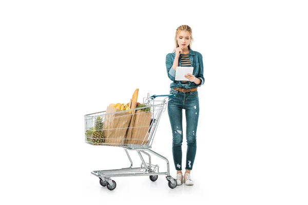 デジタル タブレットを見て 白で隔離の製品とトロリーのショッピングの近くに立っての思いやりのある女性 — ストック写真