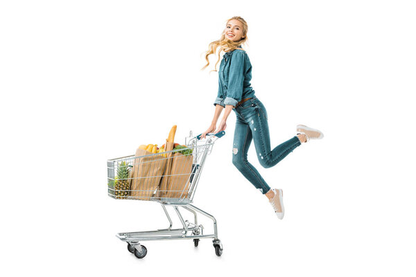 positive beautiful girl jumping near shopping trolley cart with products in paper bags isolated on white