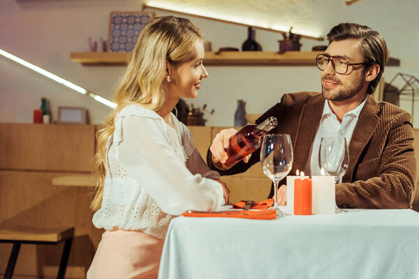 handsome man in jacket pouring wine into glass to beautiful girlfriend at table in restaurant 