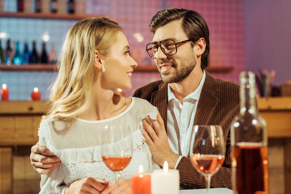 smiling man in eyeglasses and jacket embracing girlfriend at table with candles in restaurant 