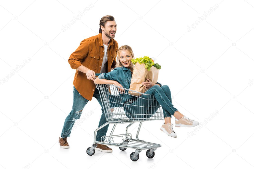 positive man carrying trolley with happy girlfriend holding shopping bags with products isolated on white