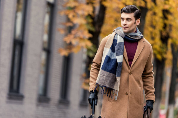 handsome stylish man in coat with umbrella walking by autumnal street