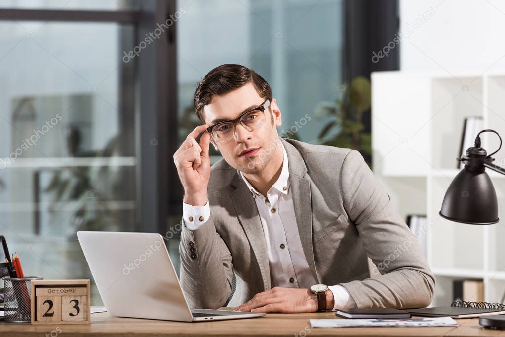 handsome businessman with eyeglasses sitting at workplace in office and looking at camera