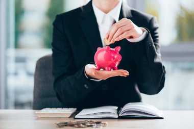 cropped shot of businesswoman putting coin into pink piggy bank 