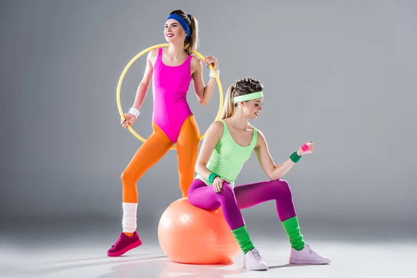 smiling sporty girls training with hula hoop, dumbbell and fit ball on grey