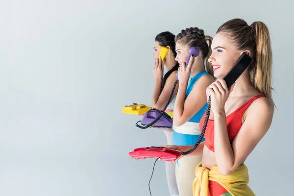 side view of smiling sporty girls talking by rotary phones and looking away isolated on grey