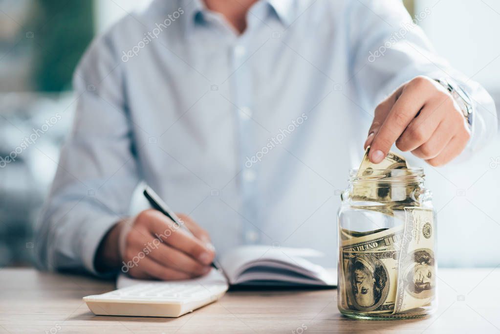 partial view of man working and putting dollar banknote in glass jar  