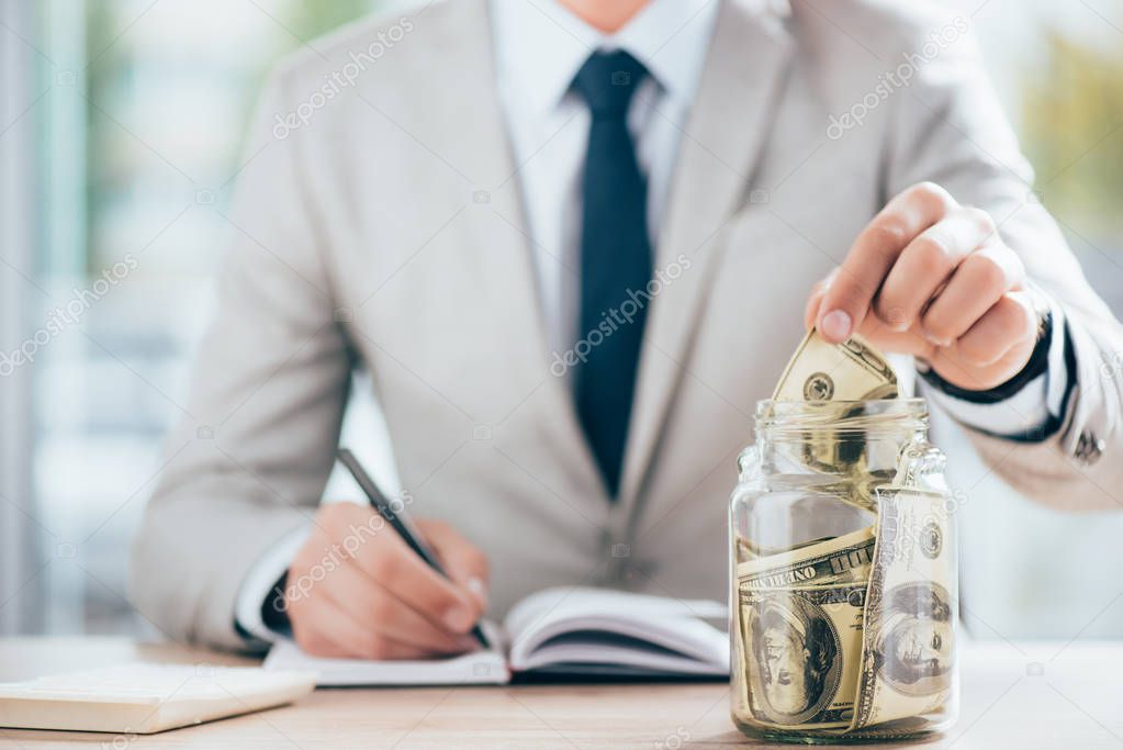 cropped shot of businessman working and putting dollar banknote in glass jar  