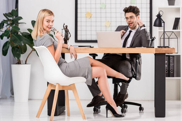 young businesswoman looking at camera while flirting with business colleague at workplace with laptop in office