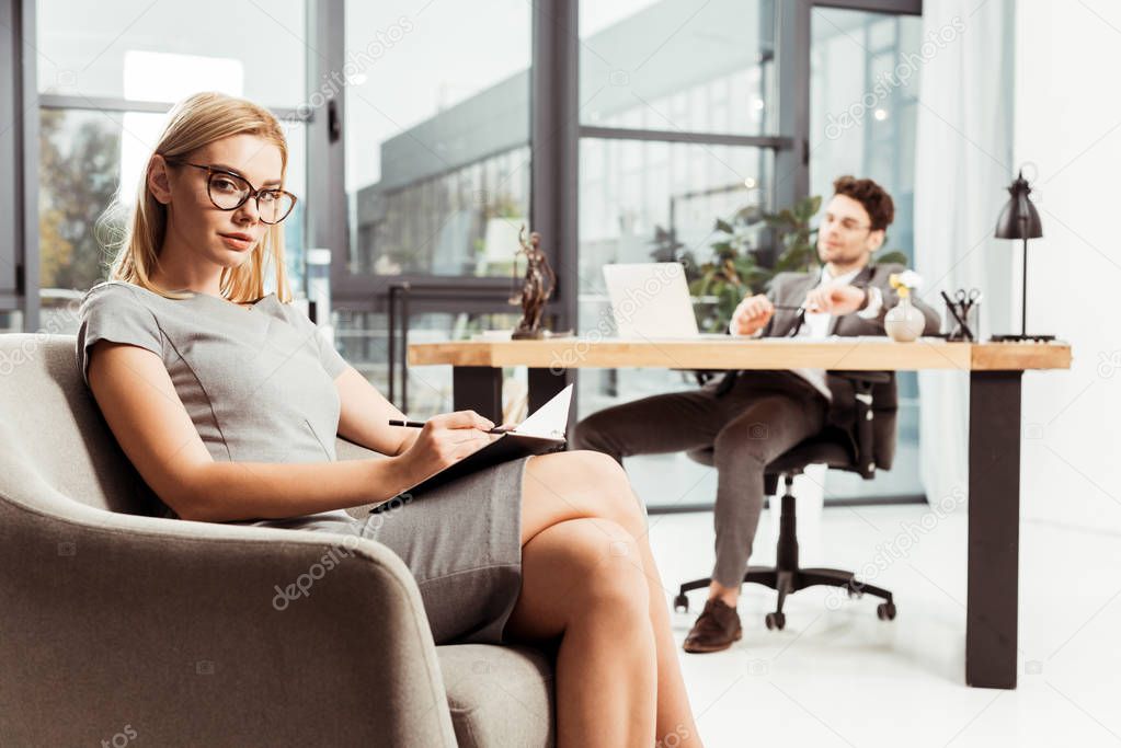 businesswoman with notebook looking at camera while sitting in armchair with colleague at workplace in office