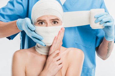 close up of plastic surgeon in gloves taping up woman scared face with bandage isolated on white clipart