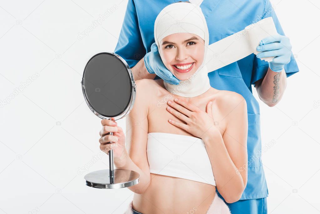 surgeon in gloves taping up face with bandage after plastic surgery while smiling woman holding mirror isolated on white