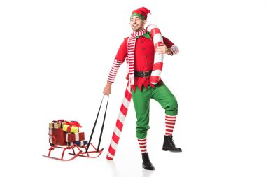 happy man in christmas elf costume carrying sleigh with presents and big candy cane isolated on white clipart
