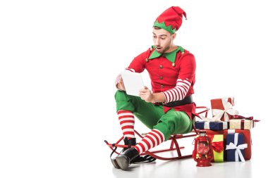 şaşırmış adam hediye yığını yakınındaki atlı kızak üzerinde oturan ve üzerinde beyaz izole wishlist bakarak Noel elf kostümü