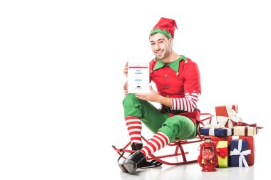 adam kızak üzerinde oturan ve üzerinde beyaz izole ekranda tablet instagram app ile tutarak Noel elf kostümü