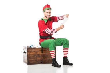 ahşap göğüs üzerinde oturan ve üzerinde beyaz izole wishlist tutan Noel elf kostümü neşeli adam