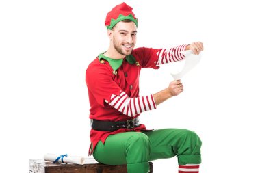 smiling man in christmas elf costume sitting and holding wishlist roll isolated on white clipart